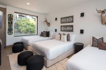 A children's bedroom at Aria Ridge featuring three single beds with white bedding, themed decor including ski signs and deer mounts, and a large window with a view of greenery.