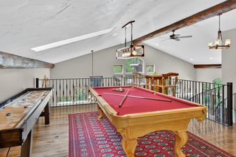 The loft at Ski Hill Sanctuary offers a games room with a red-felted pool table and a shuffleboard table, all under a vaulted ceiling with exposed wooden beams and a modern chandelier.