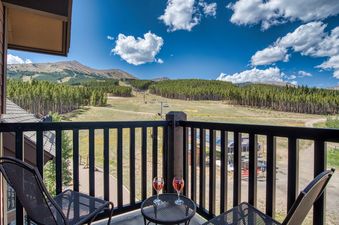 A private balcony at Crystal Peak Lodge 7407 provides a stunning view of mountains, ski slopes, and pine forests under a bright blue sky, with two glasses of rosé wine on a small table for guests to enjoy the scenery.