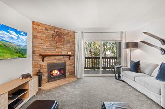A cozy living room in Snow Flower 116 Park City features a stone fireplace, a light grey sofa with blue pillows, a TV, and a balcony with an outdoor view.