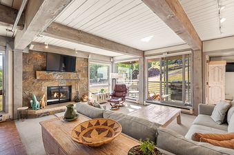 A spacious living room in Enclave 207 features a stone fireplace, a large gray sectional sofa, and a brown leather armchair, all under a ceiling with exposed wooden beams, with expansive windows looking out onto a patio and hillside.