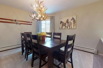 A rustic-chic dining area in Westwind 402 features a large wooden table with eight dark chairs, illuminated by an antler chandelier. Wooden skis decorate one wall, a horse painting adorns another, and a window offers mountain views.