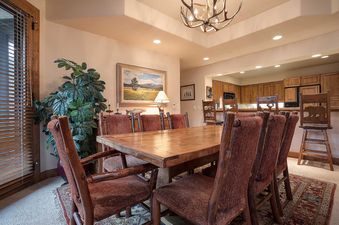 A rustic-chic dining area with a large wooden table and red upholstered chairs, featuring an antler chandelier and a large potted plant.
