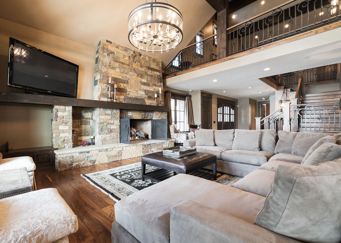 The great room at Harmony Haus features a large sectional sofa, a prominent stone fireplace with a TV above, and hardwood floors under a decorative area rug, all illuminated by a grand chandelier and overlooked by a second-story balcony.