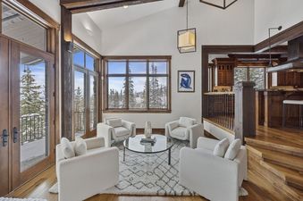 A luxurious and spacious lounge area at Silver Horse at The Colony features plush white armchairs, a glass coffee table, and large windows offering a stunning view of a snow-covered winter landscape with pine trees.