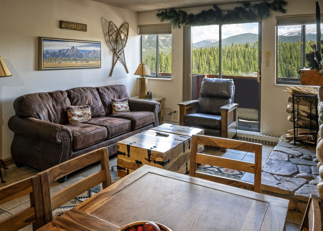 A cozy living room in the Two Bedroom Suite Beaver Run Resort with a brown leather couch, a trunk-style coffee table, and large windows looking out onto a pine forest and mountains.