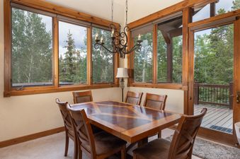 A spacious dining room at The Lodge at Cucumber Patch featuring a large wooden table and chairs, surrounded by windows offering lush green tree views.