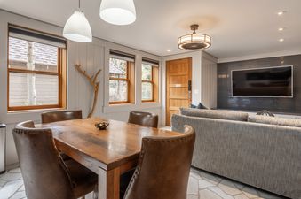 The open-concept dining area and living room at Cedar Crest Chickadee Cottage 2, featuring a wooden dining table with leather chairs and a comfortable gray sofa facing a wall-mounted TV.
