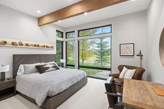 A modern bedroom with a large bed, a wooden headboard, and a long painting of bisons on the wall. The room features expansive windows offering scenic views of trees and a golf course, an armchair, and a desk.