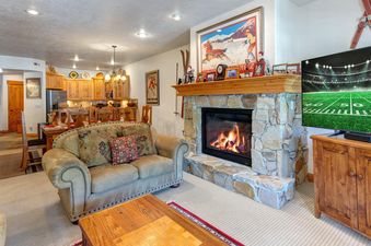 A cozy living room in Town Pointe C302 featuring a stone fireplace with a wooden mantel, a large ski-themed artwork above, a comfortable beige sofa, and a television displaying a football game.