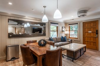 A modern and rustic dining and living area with a wooden table, leather chairs, gray sofa, and marble coffee table, with windows offering an exterior view.