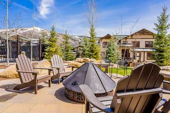 An outdoor patio area features several Adirondack chairs and a metal fire pit, with townhomes and snow-capped mountains in the background under a blue sky.