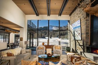 White Pine Canyon offers an open floor plan with floor to ceiling windows overlooking the snowy mountainside and ski slopes beyond. The living area has a large, comfortable couch, two plush armchairs, and a stone fireplace with a roaring fire.