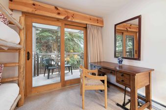 A cozy guest room features a wooden desk, a chair, and bunk beds, with sliding glass doors leading to a balcony overlooking evergreen trees.