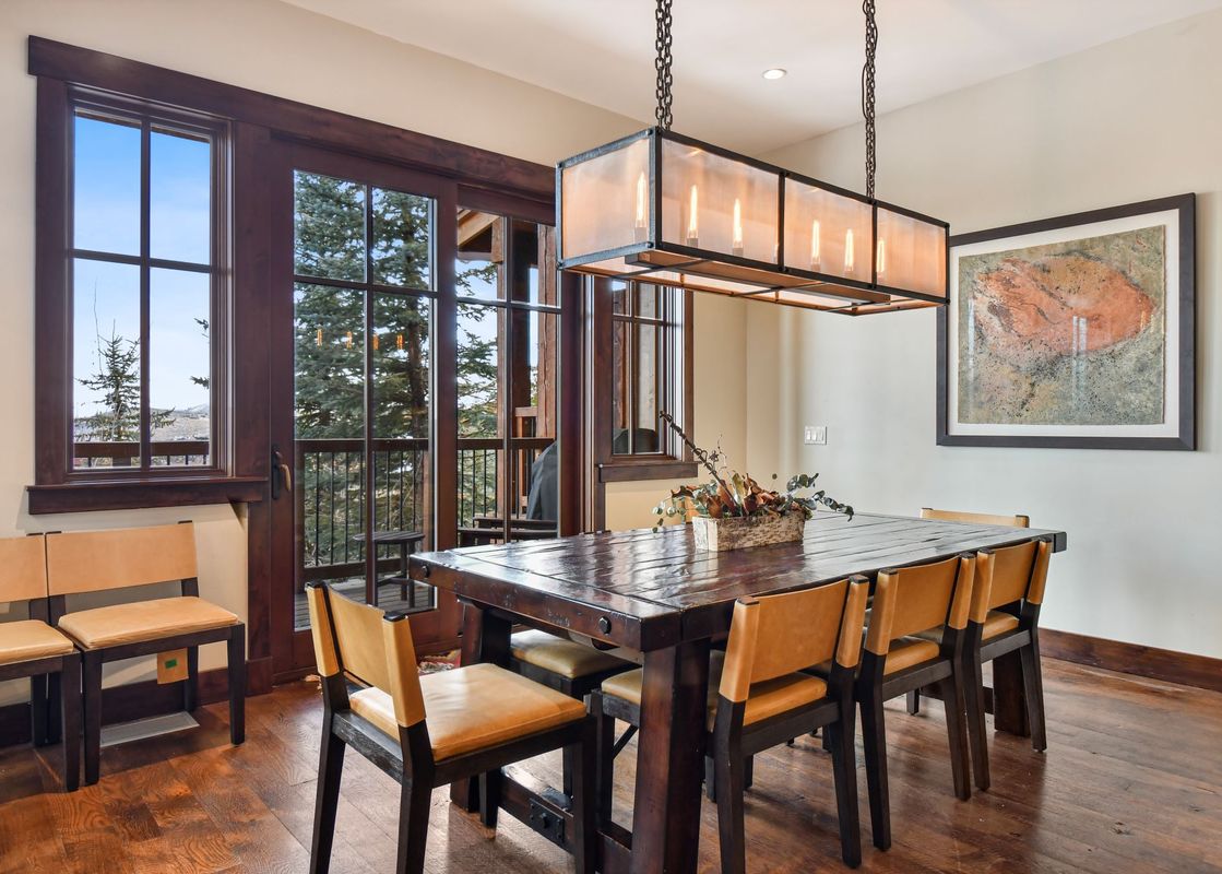 A rustic dining room with a long wooden table, leather-seated chairs, and a contemporary rectangular chandelier, featuring large windows offering views of evergreen trees and mountains.