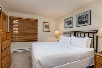 A cozy bedroom with a large bed, wooden dresser, window with blinds, and framed artwork on the walls in Montaneros 314.