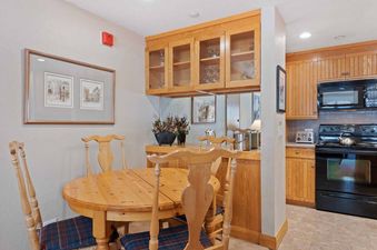 A cozy dining area featuring a round wooden table, four matching chairs with plaid cushions, and a wooden cabinet with glass doors displaying wine glasses.