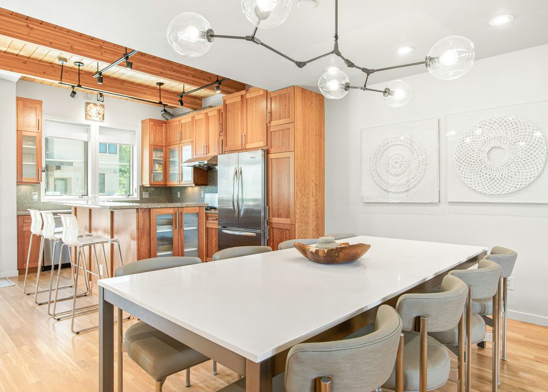 A modern dining room and kitchen with a long white table, grey chairs, wooden cabinetry, and unique lighting in Mountain Elegance 2835.