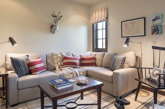 A cozy living room with an L-shaped sofa, coffee table with wine and books, two floor lamps, and various wall decorations including a deer head sculpture and framed art.