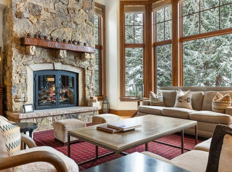 A living room with a stone fireplace, a couch, a coffee table, and a large window with a view of snow-covered trees.