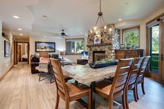 The Chateau Chamonix CX322's open-concept dining and living area features a large wooden dining table, a stone fireplace, comfortable seating, and an antler chandelier.