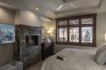 The bedroom in Three Forks Mountain One Steamboat Place 306 features a large fireplace and television, along with a bed and a large window overlooking a snowy landscape.