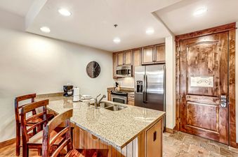 The kitchen of BlueSky Breckenridge 513 with a granite countertop and wooden cabinets, a refrigerator and microwave, and two bar stools.