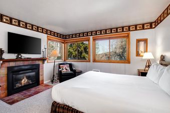 The cozy bedroom at Snow Flower 012 features a large bed, a fireplace with a TV above it, and several windows offering views of the outdoors.
