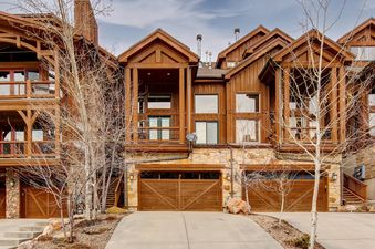 The exterior of Deer Valley 355 B, a multi-story residence with wooden siding, stone accents, multiple balconies, and two garage doors, surrounded by bare trees under a cloudy sky.