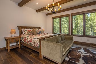 A rustic bedroom at Stony Creek Chalet features a wooden bed with a floral duvet, a cozy green sofa, and large windows looking out onto a lush forest.