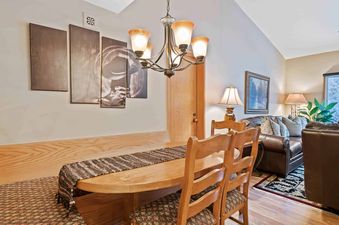 A cozy dining area at The Charter B4305 with a wooden oval table, matching chairs, and a chandelier, leading into a living space with a leather sofa.
