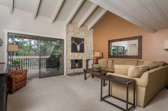 A living room with a large sectional sofa, a stone fireplace, and sliding doors that lead to a balcony with trees and a view.