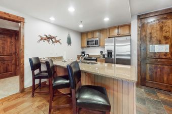 The contemporary kitchen of BlueSky Breckenridge 205 features sleek stainless steel appliances, a granite island with bar stools, and warm wooden cabinetry under recessed lighting.