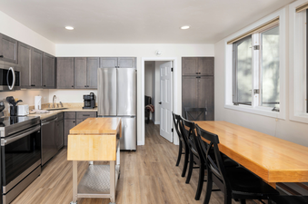 A modern kitchen with grey cabinets, stainless steel appliances, a mobile island, and an adjacent long wooden dining table with black chairs in Tamarack Townhomes 34.
