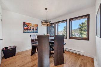 A modern dining area with a wooden table and chairs, a vibrant deer painting, and large windows offering a mountain view.