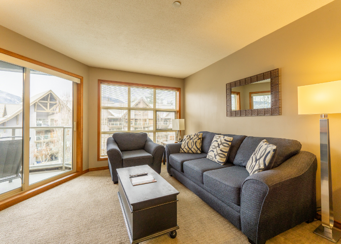 A cozy living room in Aspens on Blackcomb Unit 458 with a dark gray sofa and armchair, a black coffee table, and large windows providing natural light.