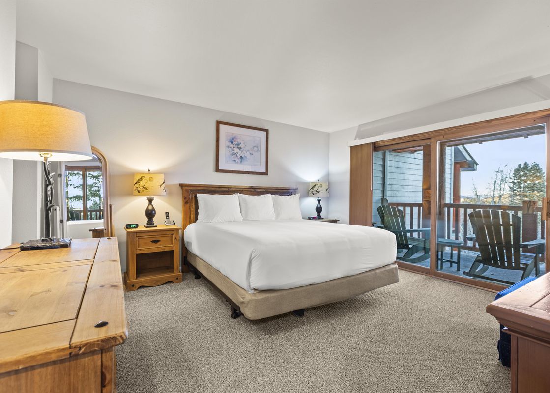 A cozy bedroom featuring a king-size bed with white linens, two wooden bedside tables with lamps, and a sliding glass door leading to a balcony with wooden chairs and a view of the outdoors.