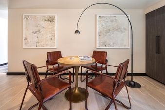 A game table with four brown leather chairs and a puzzle box, illuminated by a modern black arched floor lamp, set against light walls with abstract art.