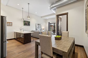 The dining area at 2nd Street Lofts 209 features a rustic wooden table and upholstered chairs, seamlessly connecting to the living room with a sectional sofa and a patio door leading to an exterior view.