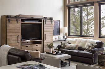 A rustic, cozy living room at The Nest in Jackson Hole with leather sofas, faux-fur throws, a large wooden entertainment center, and scenic forest views.