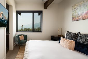 A bedroom with a large window overlooking the mountains, a bed with white linens and pillows, a green armchair and a nightstand.
