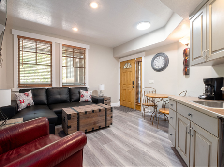 The living area of Town Pointe C106 features a black leather sectional sofa, a rustic wooden chest coffee table, and a red armchair, with a kitchen area and dining table visible beyond.