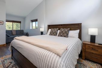 The comfortable bedroom in Vasquez Creek Townhomes 8 features a large bed with a dark wood headboard, striped bedding, and patterned pillows, with a sofa and windows visible in the background.