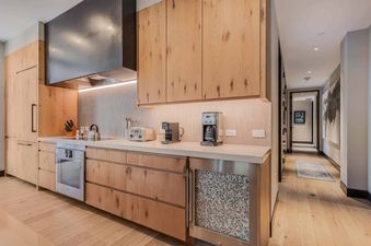 A modern kitchen in One Snowmass East 704 featuring light wooden cabinetry, white countertops, and stainless steel appliances, with a hallway visible to the right.