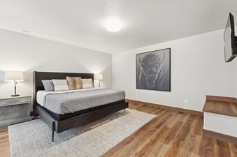 A master bedroom featuring a large bed with a dark headboard, two nightstands with lamps, a prominent bison artwork on the wall, and a mounted television, all set against light walls and a wooden floor.