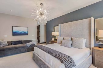A modern bedroom with a large bed, upholstered headboard, chaise lounge, and a starburst chandelier, featuring blue accent walls and mountain artwork.