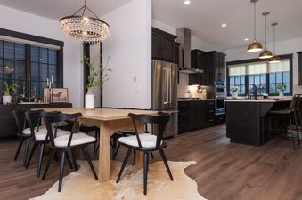 A dining area with a rustic wooden table, black chairs with white cushions, and a crystal chandelier, adjacent to a modern kitchen with dark cabinetry and stainless steel appliances.