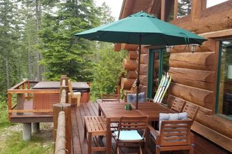 The Hellroaring Hut features a large wooden deck with a hot tub, an outdoor dining set under a green umbrella, and a view of surrounding pine trees.