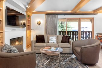 A cozy living room in the Bridge Street Lodge 1 Bedroom Gold Unit 308, featuring a television, a lit fireplace, comfortable seating, and access to a balcony with an outdoor view.
