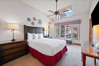 A master bedroom at The Charter F4405, featuring a large bed with white bedding and a red bed skirt, dark wood furniture, snowflake wall art, and windows overlooking a mountain landscape.